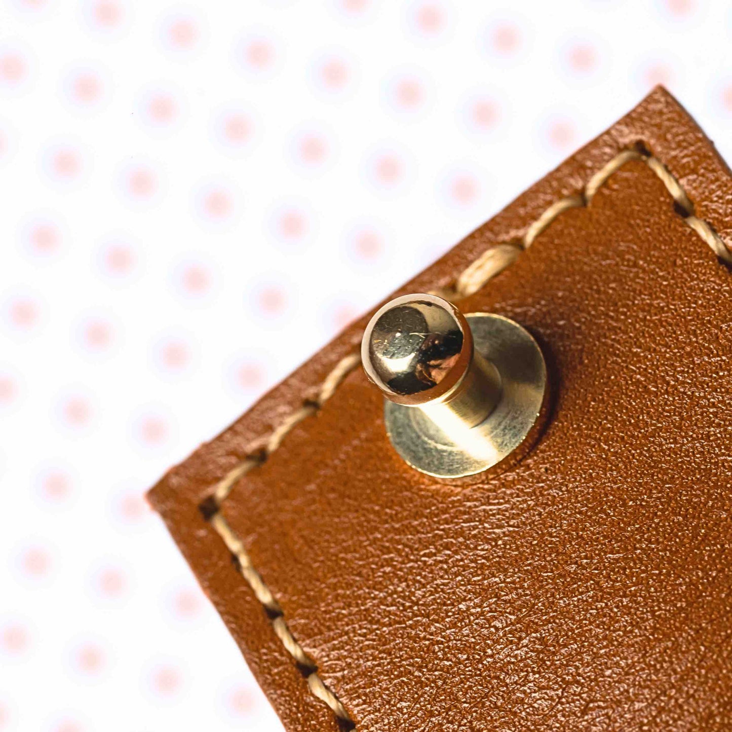 Close ingezoomd beeld van goudkleurig sluitingsknopje van bevestigingssysteem van eco leren cover haaraccessoire in kleur cognac met witte stik Achtergrond is blurry wit met oranje puntjes.
