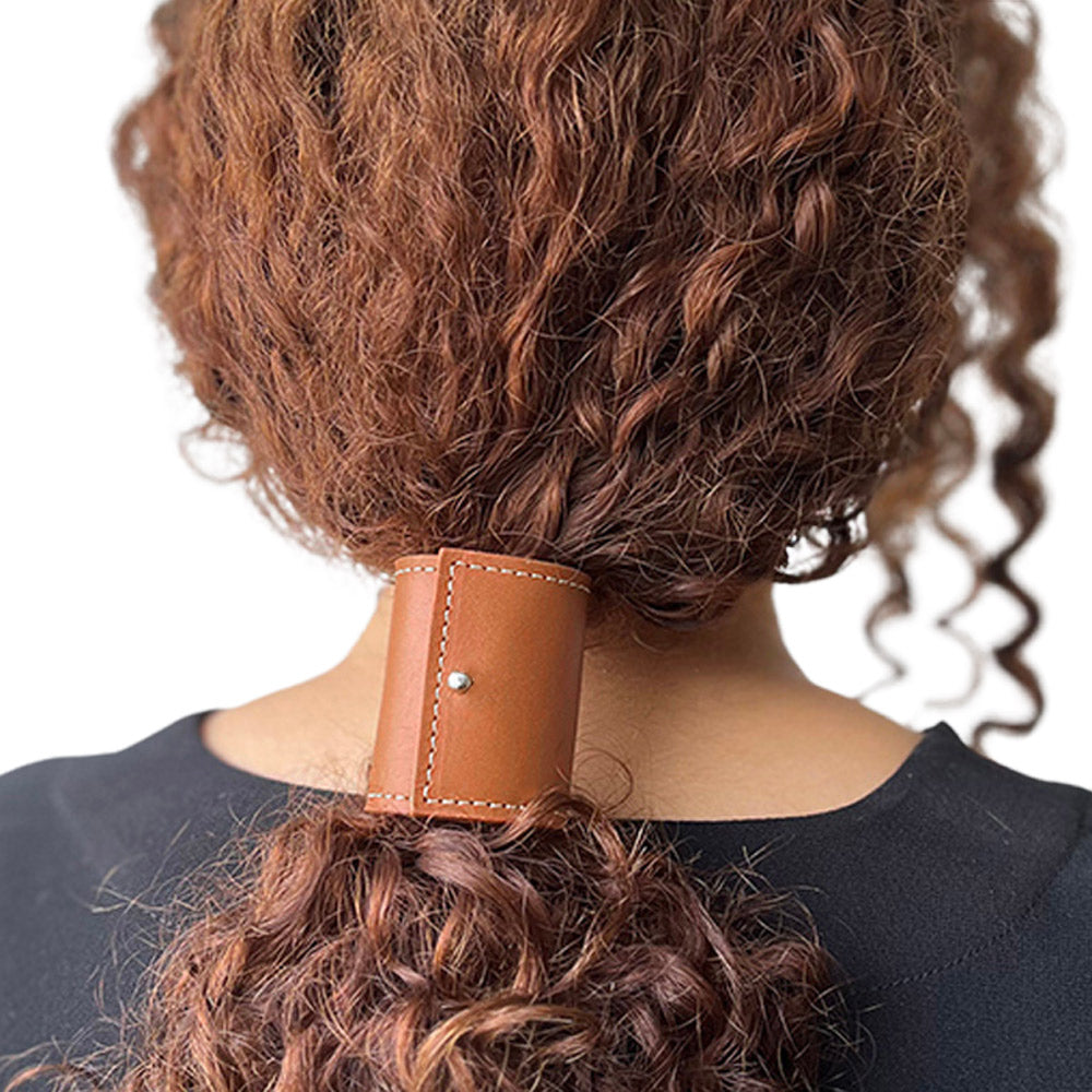 Model met krullend bruin lang haar, losjes vastgezet met een lange leren band in kleur cognac, maat L met gouden sluitingsknopje in kleur cognac bruin