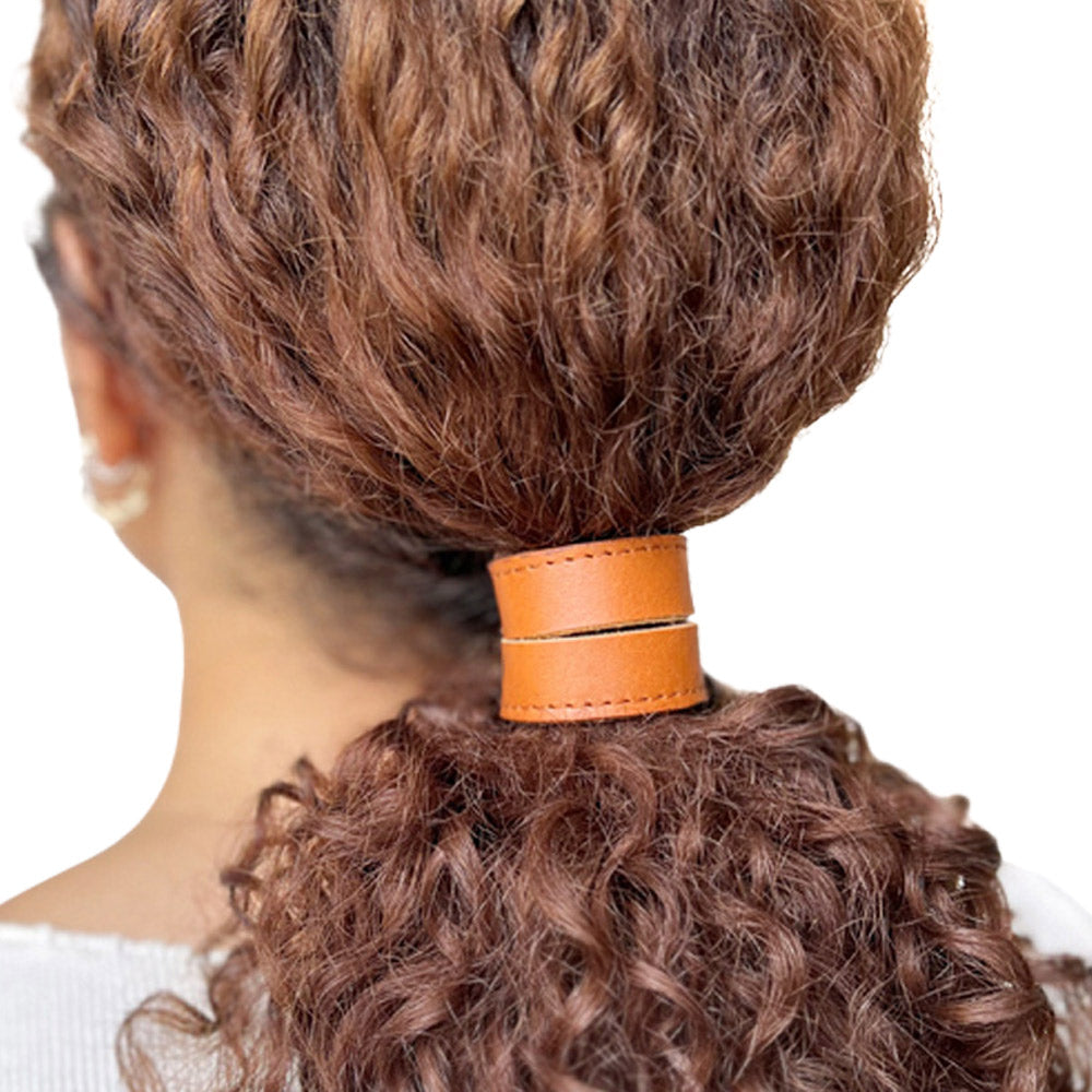 Model met dik krullend kastanjebruin haar, in een losse lage staart vastgezet met leren 2-band haarelastiek in kleur cognac. Het model draagt een maat L.