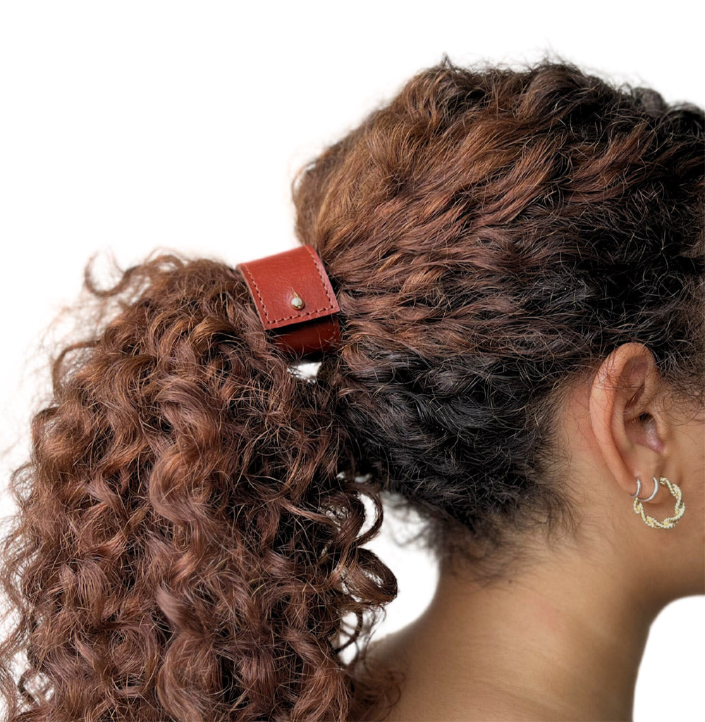 Model met dik krullend kastanjebruin haar. Ze draagt het in een hoge paardenstraat en het is vastgezet met korte leren haarban in de kleur Sienna bruin in maar L.