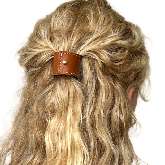 Model met golvend blond lang haar, haar is bovenop, alleen de bovenlaag van het haar, vastgezet met een korte leren haarband in de kleur cognac met witte stiksels. Het model draagt een maat S